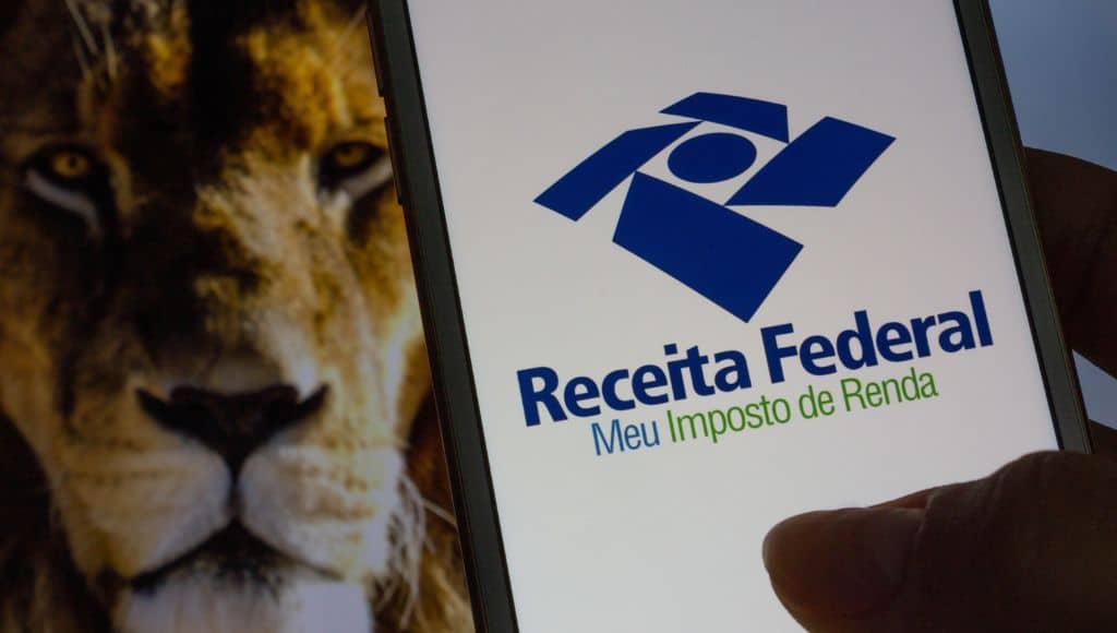 imagem de um celular com o site da receita federal aberto. ao fundo há a imagem de um leão