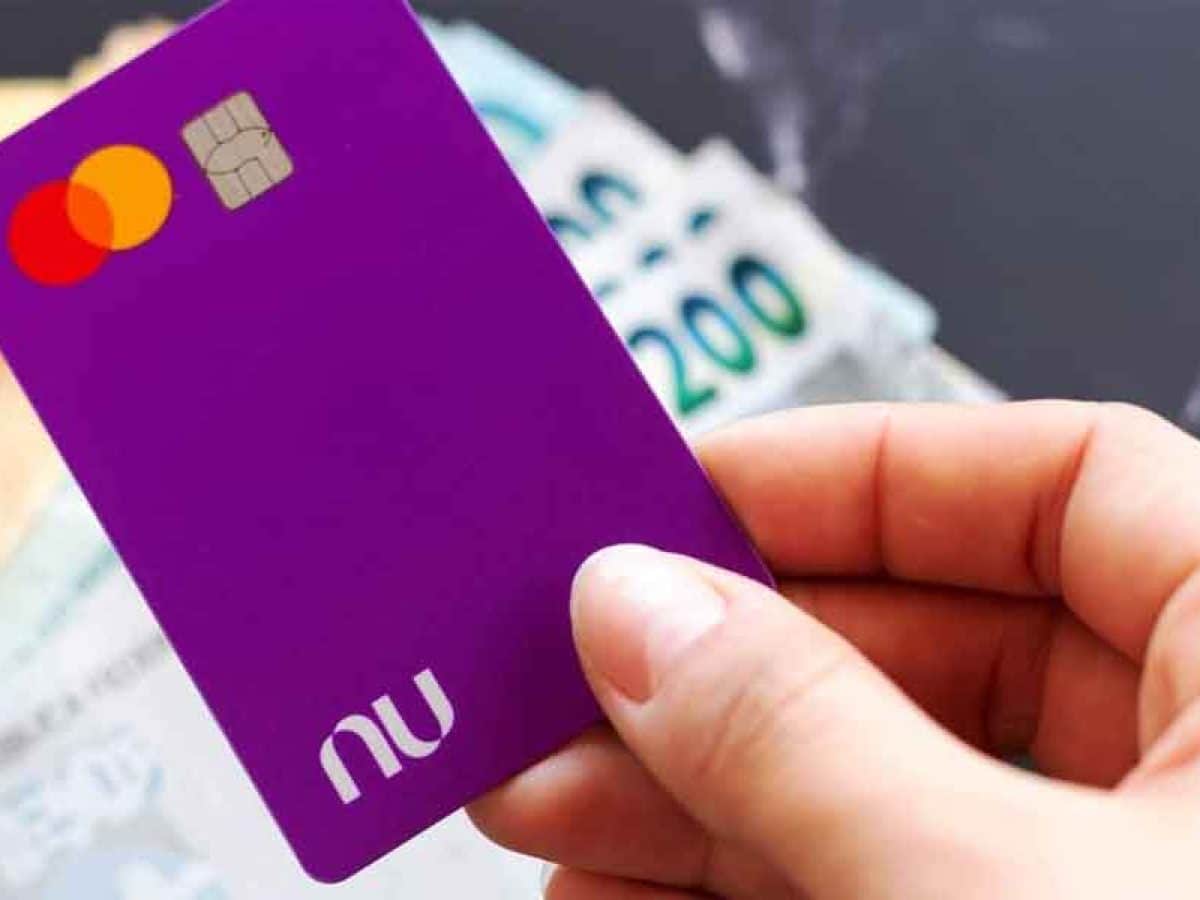 Mão feminina segurando cartão roxo do Nubank e ao fundo se vê notas de 200 reais.
