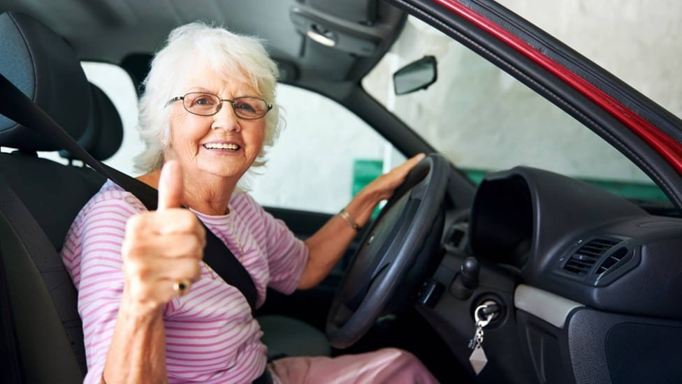 Mulher idosa, com cabelos totalmente brancos, usando óculos e roupa cor de rosa, ao volante