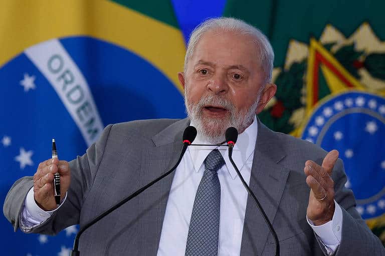 Foto do presidente Lula falando ao microfone, atrás dele uma bandeira do Brasil e uma da bandeira presidencial.
