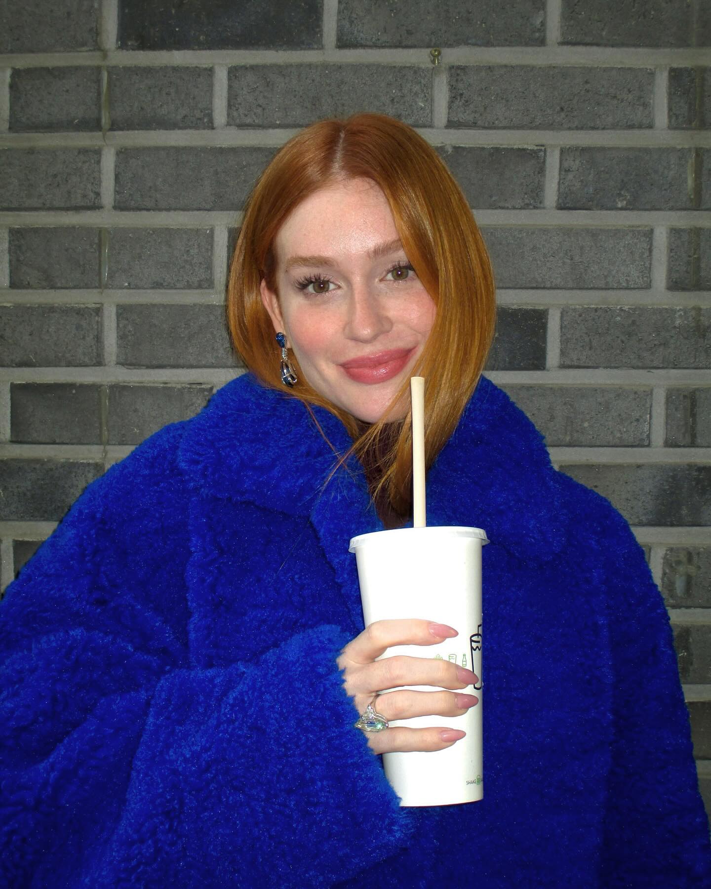 Marina posa para a foto, ela esta usando casaco de pelo azul, segurando um copo branco.