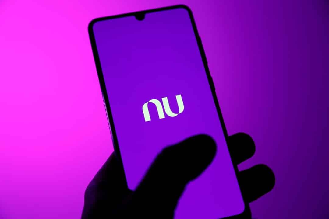 mão segurando smartphone com logo do nubank. fundo da foto é roxo.