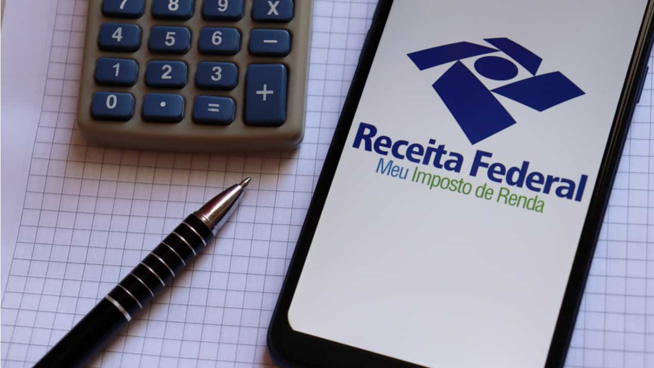 celular com aplicativo da receita federal aberto sobre uma mesa coberta com papel quadriculado ao lado de uma lapiseira e uma calculadora