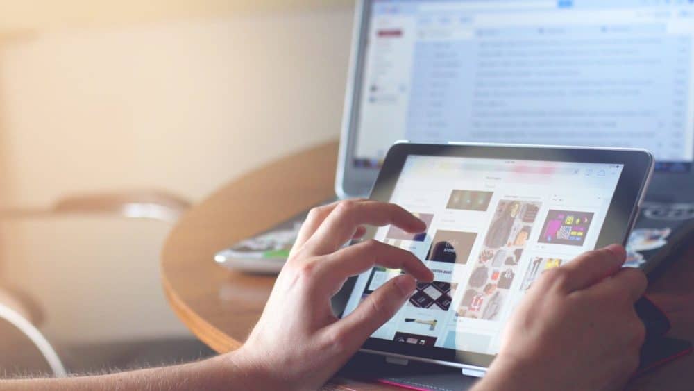 imagem de uma pessoa com um computador ao fundo e um tablet mão fazendo compras on-line para falar sobre a taxação e-commerce