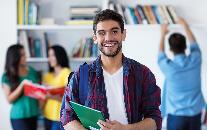 jovem branco sorrindo segurando caderno, ao fundo há duas moças e um rapaz.