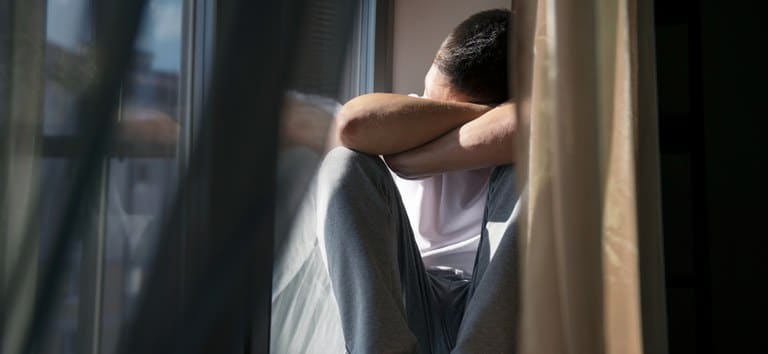 pessoa sentada com braços e cabeça apoiados sobre os joelhos ao lado de uma janela fechada.