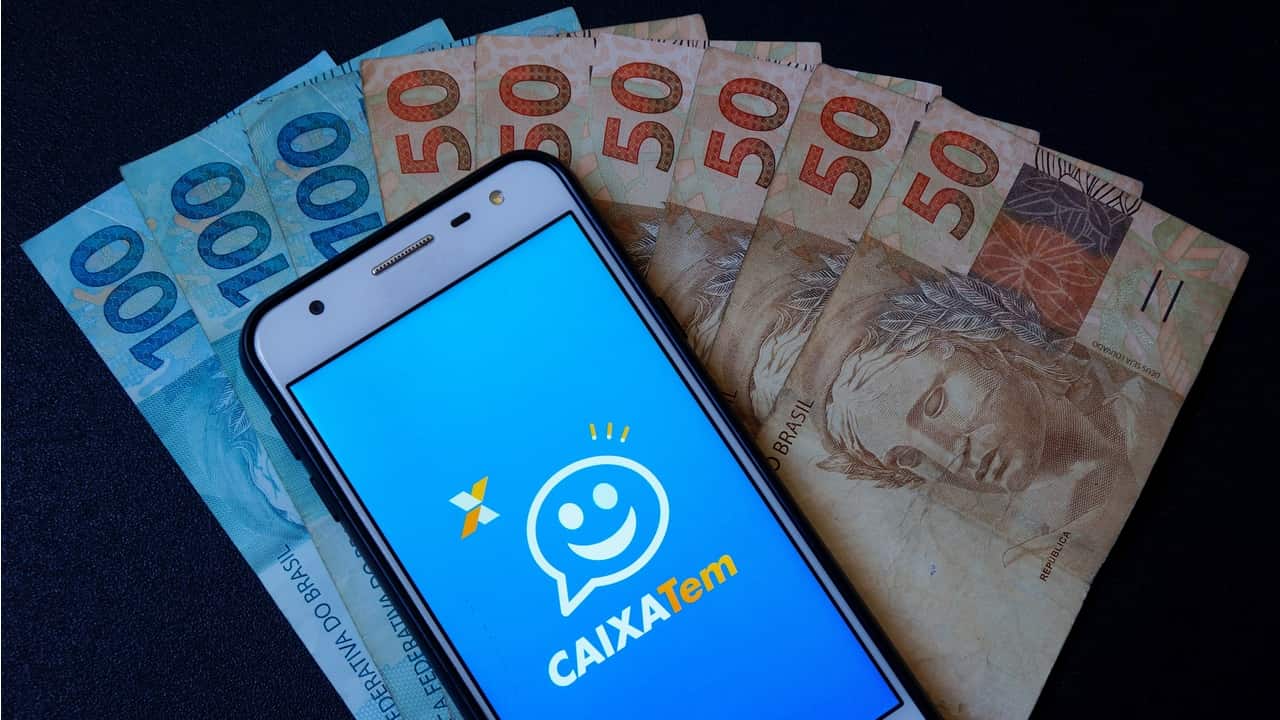 notas de 100 e 50 reais. celular com app do caixa tem aberto. (Fonte: Reprodução Remessa Online)