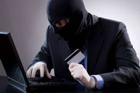 homem usando capuz preto, terno e luvas, segura um cartão magnetico e usa um laptop.