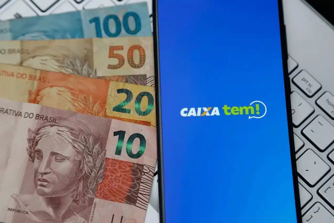 celular aberto no aplicativo da caixa e ao lado de dinheiro