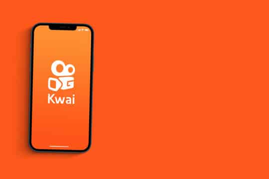 smartphone com aplicativo kwai aberto sobre um fundo laranja. (Fonte: Reprodução Adobe Stock)