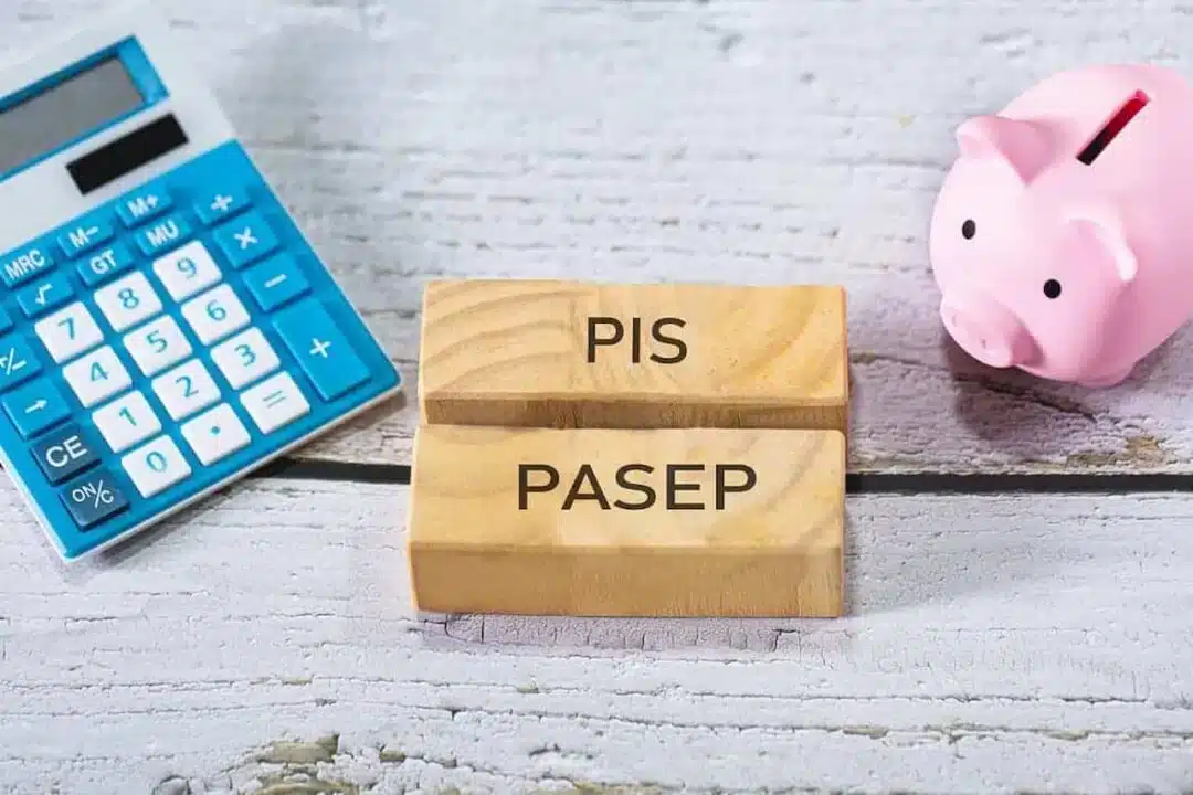 blocos de madeira com 'pis pasep' escrito, sobre tábuas de madeira, entre uma calculadora azul e porquinho (cofre) rosa