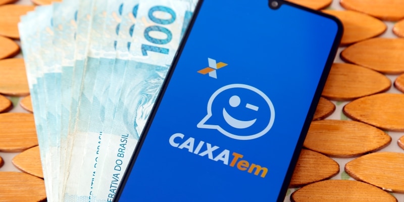 smartphone com app do caixa tem aberto e notas de 100 reais ao lado